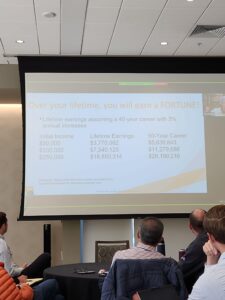 Audience members at the NAIFA Utah Symposium watch a presentation slide discussing lifetime earnings, long-term wealth building, and financial wellness during a retirement planning session.